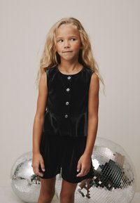 Crni plišani top bez rukava s gumbima i peplum dizajnom, uparен s odgovarajućim kratkim hlačama. U pozadini se vide disco kugle s ogledalima.