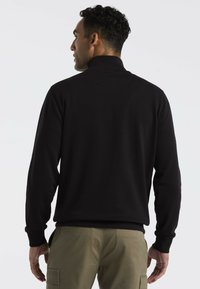 Schwarze Zip-Sweatjacke mit hohem Kragen. Rippbündchen und Saum. Getragen mit hellen Khakihosen, die seitliche Taschen und Cargo-Stil aufweisen.