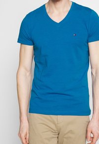 Blaues V-Ausschnitt-T-Shirt aus Baumwolle mit kleinem Logo auf der Brust. Kurze Ärmel und taillierte Passform. Glatte Textur.