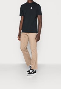 T-shirt en coton noir avec logo blanc, associé à un pantalon en coton beige ; des baskets noires et blanches complètent la tenue.