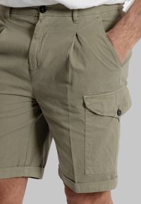 Pantaloni cargo color khaki con tasche laterali, pieghe frontali, chiusura a bottone e orlo arrotolato. Realizzati in un tessuto leggero, presentano una consistenza liscia.