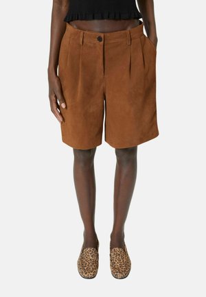 Person iført brune shorts med høj talje og plissering samt loafers med leopardprint, stående med den ene hånd i lommen mod en hvid baggrund.