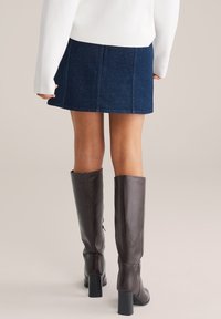 Sötétkék farmer mini szoknya láthat�ó varrásokkal, párosítva térd fölé érő barna bőr csizmával, amely négyzetes sarkú és cipzáros részletekkel rendelkezik.