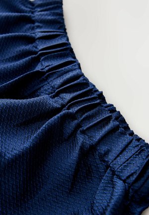 Tmavomodrá látka s textúrovaným diamantovým vzorom, s nariasenými volánmi pozdĺž jedného okraja, ktoré vytvárajú hĺbku a dimenziu.