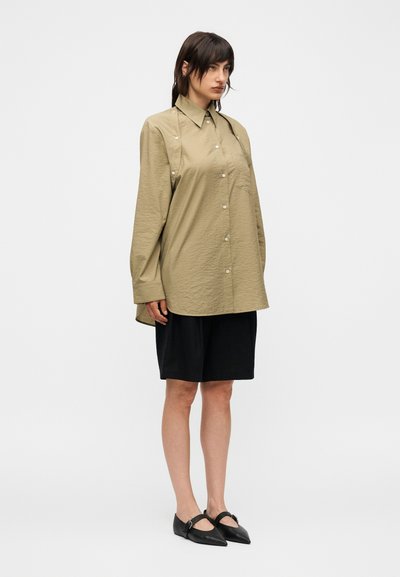 Γυναίκα που φορά υπερμεγέθη μπεζ πουκάμισο με κουμπιά, μαύρη φούστα μέχρι το γόνατο και μαύρα μυτερά φλατ παπούτσια, στέκεται μπροστά σε απλό φόντο.