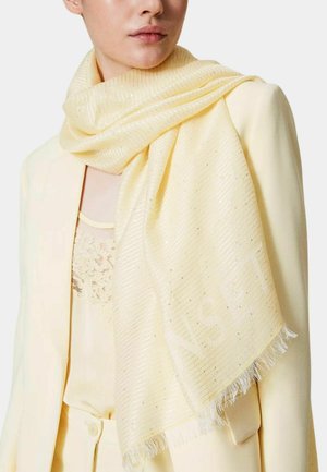 Foulard giallo chiaro con strisce testurizzate e luccichio, bordi frangiati, appoggiato su una giacca e una maglia di colore chiaro abbinate.