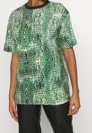 Personne portant une chemise à manches courtes verte et noire à motifs, col noir, et un pantalon noir sur un fond blanc uni.