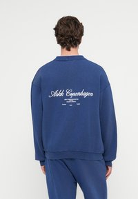 Sötétkékkék kapucnis pulóver laza fazonnal, gallérral, bordás mandzsettákkal, és fehér szöveges hátsó nyomattal. Puha, texturált anyag.