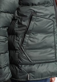 Zelená péřová bunda s texturovaným povrchem, která má diagonální zipovou kapsu na levé straně a značkovou visačku na spodním lemu.