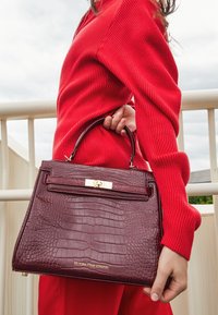 Burgunderfärgad handväska med krokodilmönster och en strukturerad form, guldtonad hårdvara och ett handtag upptill, som hålls vid sidan av en person i en röd ribbad tröja.