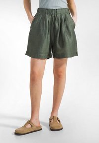 Pantaloncini verdi in lino con una fascia larga in vita, pieghe e tasche. Abbinati a scarpe slip-on beige in camoscio con dettagli a taglio.