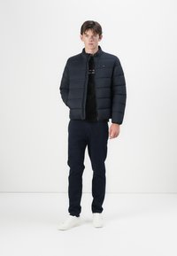 Marineblå pufferjakke med høyt krage, quiltet design og glidelås. Båret over en svart t-skjorte og parret med marineblå bukser og hvite sneakers.
