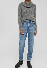 Femme portant un pull turtleneck en tricot épais gris, un jean taille haute bleue clair avec un revers, et des bottes noires à lacets.