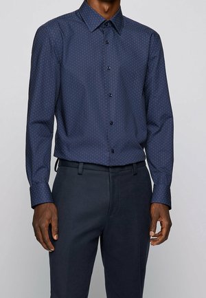 Mann trägt ein marineblaues Hemd mit kleinen roten Punkten, das in passende marineblaue Hosen gesteckt ist, vor einem einfarbigen Hintergrund.