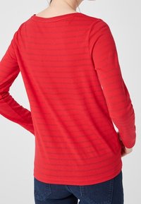 Maglia rossa a maniche lunghe con sottili righe scure, scollatura rotonda e silhouette aderente, abbinata a jeans blu.