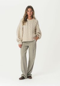 Cremfarbener Grobstrickpullover mit übergroßen Ärmeln, kombiniert mit hellgrauen Hosen im taillierten Schnitt. Braune Schuhe runden das Outfit ab.