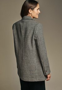 Pilkas ir smėlinis languotas švarkas su struktūruotais pečiais, susegamomis rankogaliais ir šiek tiek ilgesne nugarėle, pagamintas iš tekstūruotos medžiagos.