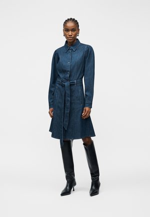 Kobieta ubrana w długą, zapinaną na guziki sukienkę denimową z paskiem w talii, w połączeniu z czarnymi, sięgającymi do kolan kozakami na obcasie, stojąca na jednolitym tle.