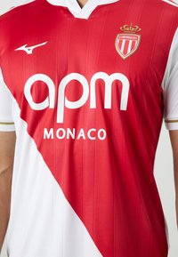 Punainen ja valkoinen urheilupaita pystysuorilla raidoilla, jossa on V-pääntie, logo vasemmassa rinnassa ja suurilla valkoisilla kirjaimilla teksti "apm MONACO."