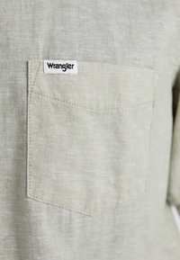 Zelená ľanová košeľa s krátkym rukávom, ktorá má náprsné vrecko s bielou nálepkou "Wrangler" a jemnú štruktúru.