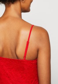Camisola rossa in pizzo con spalline sottili regolabili, che presenta motivi floreali e una texture liscia. Il design dettagliato sul retro mette in risalto la ricchezza del tessuto.