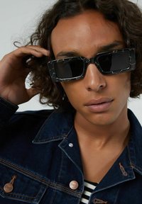 Lunettes de soleil noires avec des montures rectangulaires, bords épais à motifs, portées avec une veste en jean foncée et une chemise rayée noir et blanc.