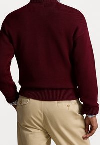 Burgundy stickad tröja med ribbat mönster, hög hals och avslappnad passform, kombinerad med beiga byxor som har en bakficka och dragkedjedetalj.