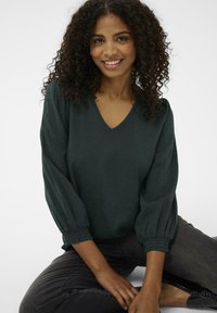 Donkergroene, textuur blouse met een V-hals en opbollende mouwen, voorzien van gegatherde manchetten en een relaxte pasvorm, gecombineerd met donkergrijze jeans.