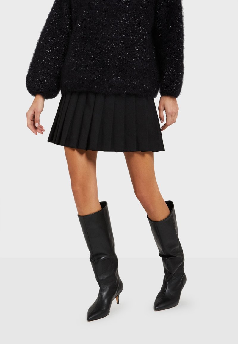 Zwarte geplooide rok met een gestructureerde, fluffy zwarte trui en zwarte leren kniehoge laarzen met puntige neuzen en hoge hakken.