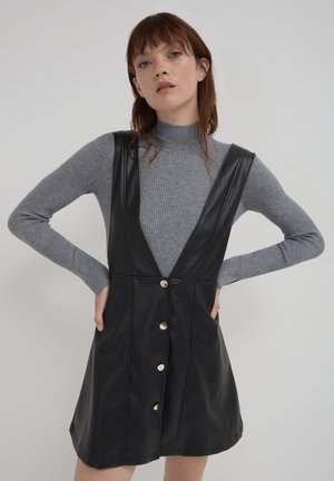Robe sans manches en faux cuir noir avec un décolleté en V profond, des accents en boutons dorés et une silhouette ajustée. Superposée à un col roulé gris.
