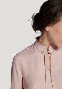Langärmlige Bluse aus zartem pinken Stoff mit hohem Kragen, Schlitzöffnung und einer Schleifendetail. Sanfte Textur und subtiler Glanz.