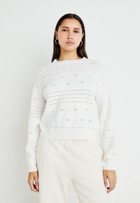 Pulover alb din tricot cu model din dantelă, guler rotund și manșete ripsate. Textură moale, croială lejeră, asortat cu pantaloni de culoare deschisă.