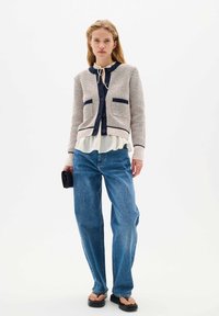 Knit cardigan in cream with navy stripes, featuring two front pockets, a collar, and buttons, layered over a white blouse and paired with blue jeans.