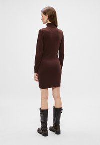Vestido de punto marrón chocolate con cuello alto, silueta ajustada y mangas largas. Combinado con botas negras hasta la rodilla con hebillas.