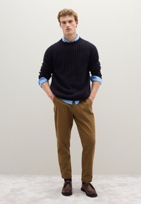 Marineblauer Rippstrickpullover, darunter hellblaues Hemd mit Kragen, beigefarbene Hose und braune Schuhe. Einfaches, lässiges Outfit mit geschichteten Texturen.