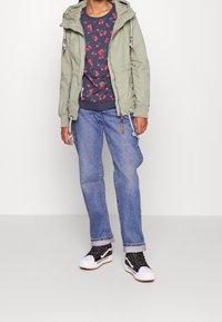 Ljusgrön hoodie med dragsnören, marinblå skjorta med röd körsbärsmönster, blå jeans och svarta sneakers med vita sulor.