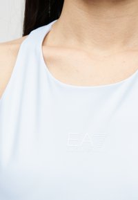 Γυναίκα που φοράει αμάνικη γαλάζια μπλούζα με μικρό ανάγλυφο λογότυπο EA Emporio Armani στο στήθος.