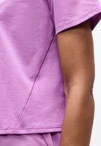 Κοντινό πλάνο βραχίονα ατόμου που φοράει πουκάμισο με κοντό μανίκι λεβάντας και παντελόνι που ταιριάζει, δείχνοντας την υφή του υφάσματος και τις λεπτομέρειες των ραφών.