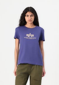 Violet medvilninis marškinėlis su trumpomis rankovėmis ir apvaliu iškirpimu, su smėlio spalvos logotipo spauda, kurioje yra tekstas "ALPHA INDUSTRIES."