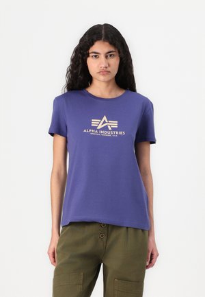 Camiseta de algodón morada con mangas cortas y cuello redondo, que presenta una impresión de logo beige que incluye el texto "ALPHA INDUSTRIES."