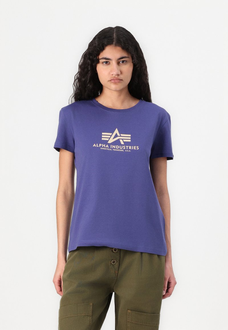 Violet medvilninis marškinėlis su trumpomis rankovėmis ir apvaliu iškirpimu, su smėlio spalvos logotipo spauda, kurioje yra tekstas "ALPHA INDUSTRIES."