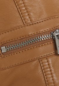 Hnědý syntetický kožený materiál s viditelným švem. Stříbrný kovový zip probíhá podél švu a ukazuje texturovaný materiál na rozhraní.