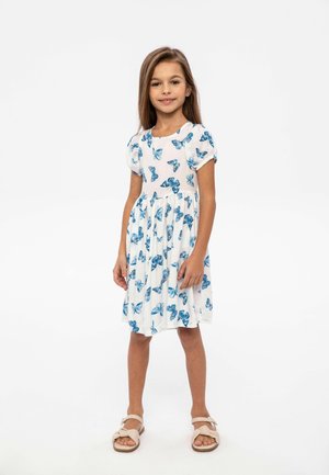 Robe blanche avec imprimé papillon bleu, manches courtes bouffantes, taille cintrée et jupe évasée. Associée à des sandales beiges.