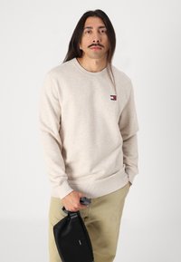 Beige bomullsgenser med lange ermer og ribbestrikkede mansjetter, med en liten logo-patch på brystet. Modellen holder en svart veske.
