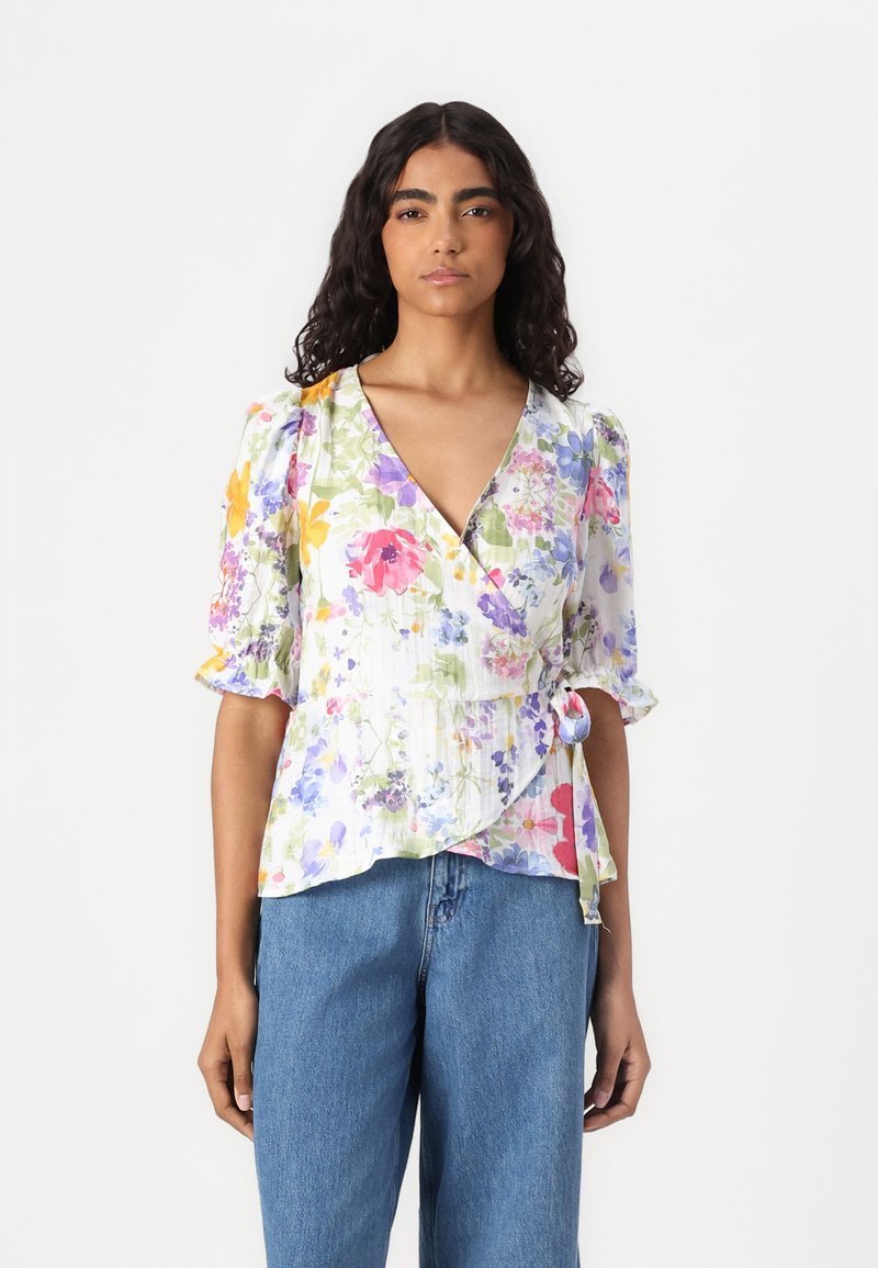Blomstrete innpakningsbluse i lett stoff med puff-ermar, prydet med en blanding av lilla, gule og rosa blomster på hvit bakgrunn. Knytt detalj på siden.