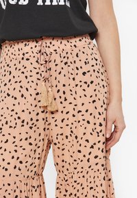 Világos rózsaszín nadrág, fekete pöttyös mintával, húzózsinóros derékkal és bojtokkal a végén. Az anyag lágy és lebegő hatású.