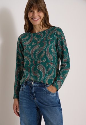 Femme souriante portant un haut à manches longues vert à motif paisley et un jean bleu, une main dans la poche, sur un fond uni.