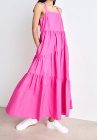 Różowa, warstwowa sukienka maxi z cienkimi ramiączkami i kieszeniami, wykonana z lekkiego materiału. Połączona z białymi trampkami.