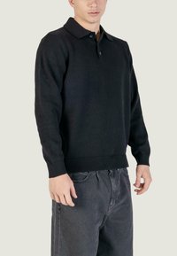 Maglione polo nero a maglia con colletto, due bottoni, polsini e orlo a coste. Indossato con pantaloni grigio scuro, con una vestibilità comoda.