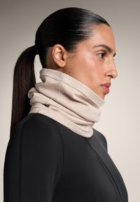 Világos bézs, puha, struktúrált nyakmelegítő, hurkolt anyagból, fekete felső felett viselve. Sima, testhezálló kialakítás rétegzett redőkkel.